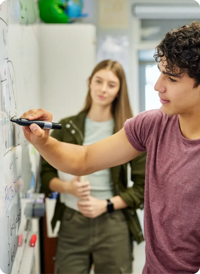 Student writing on a whiteboard with another student in the background.